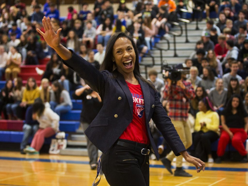 Keri Pilgrim-Ricker smiling and waving as she addresses a gymnasium of students.