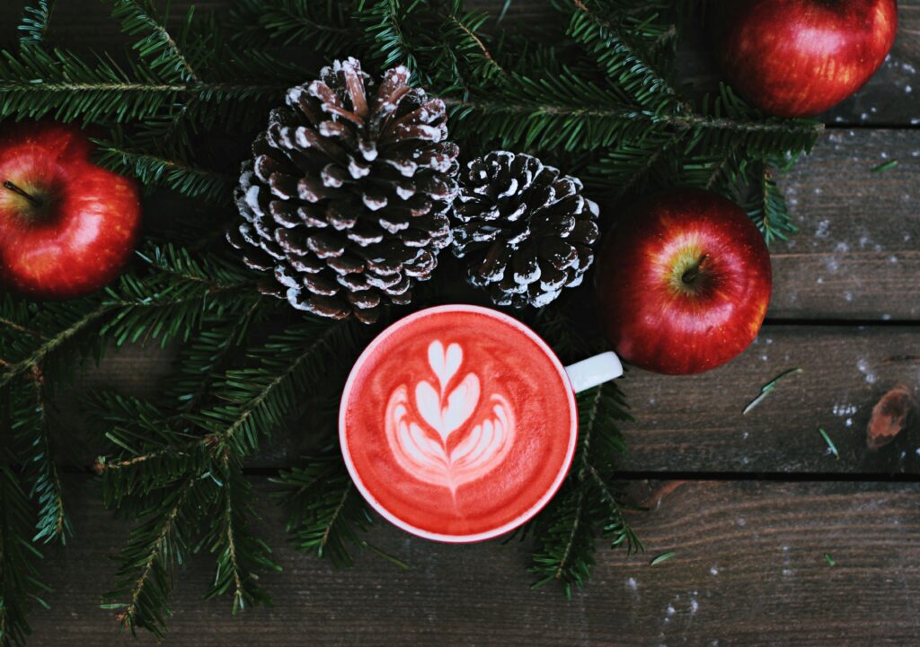 Cup of coffee with design of hearts in the milk foam, surrounded by apples, fir boughs and white frosted pine cones.