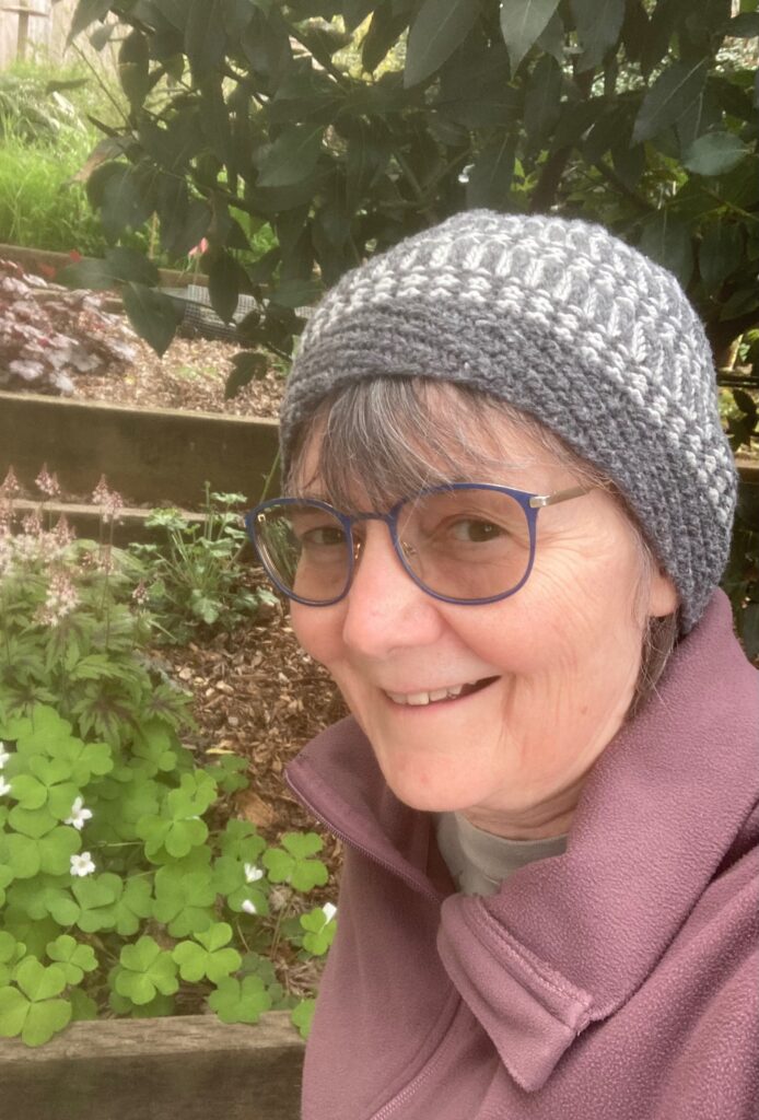Karen Anderson, smiling, wearing a cap, and standing in front of green plants.