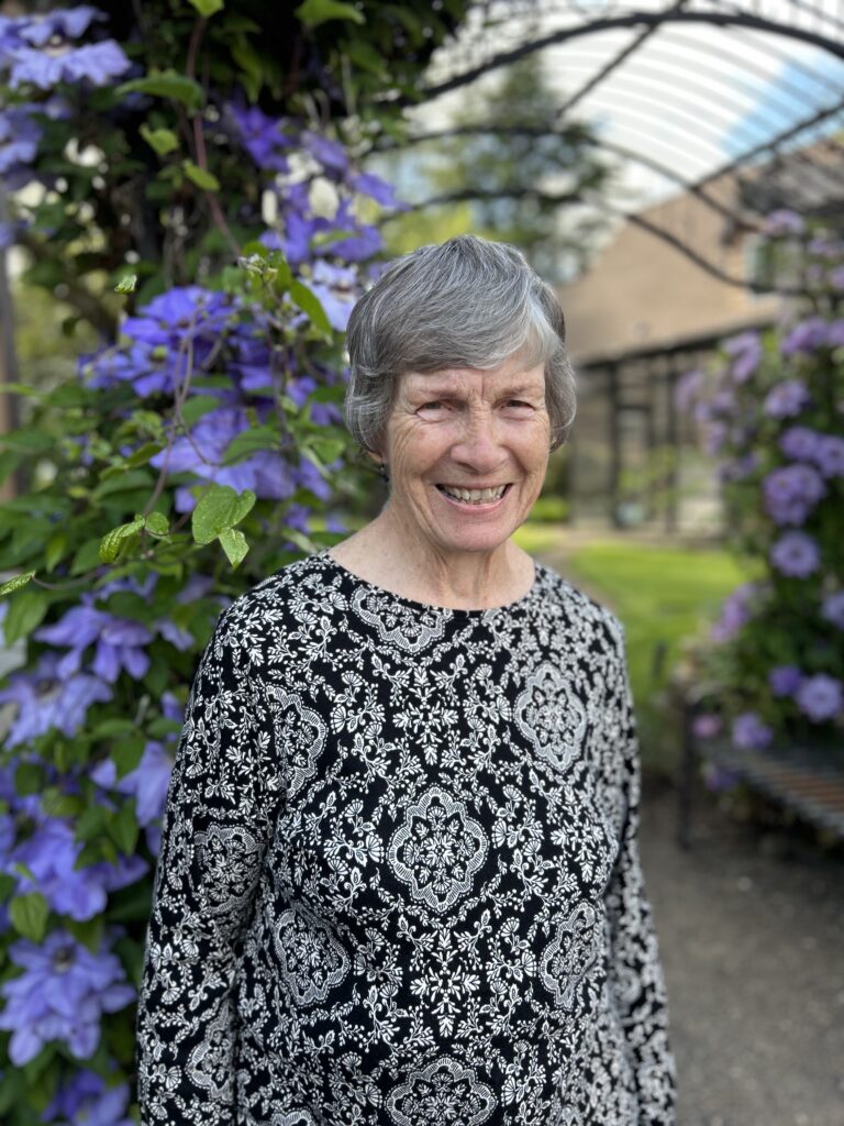 Nancy Muir, smiling and standing in front of blooming purple clematis flowers.