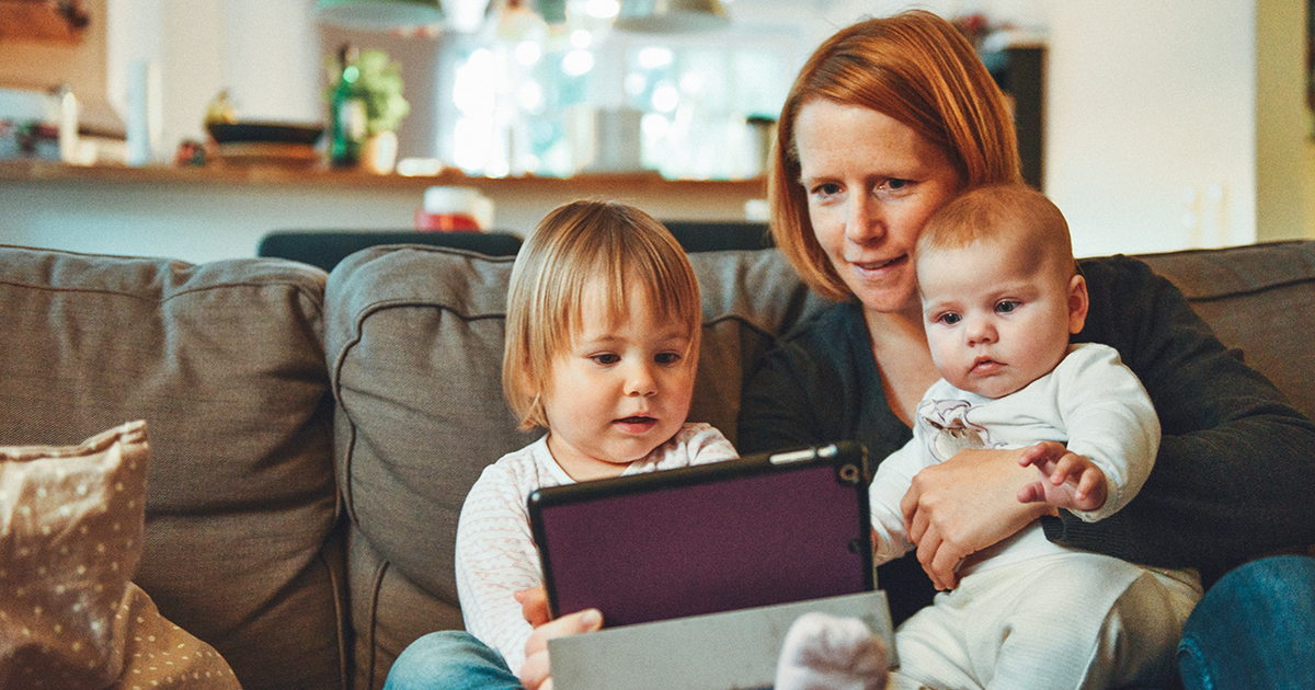 mom, toddler and a baby sit on a couch while viewing an iPad