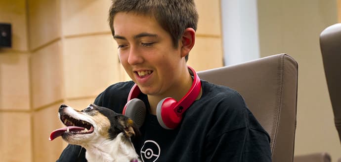 young boy sits with dog in lap, headphones around his neck and smiling, dog has his tongue out