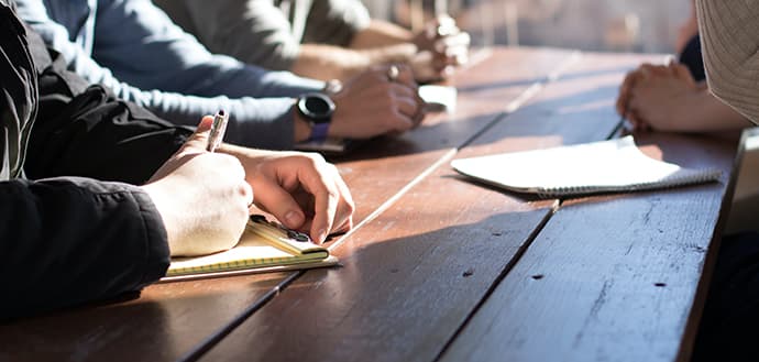 four people sit at desk, writing on notepads, no faces shown