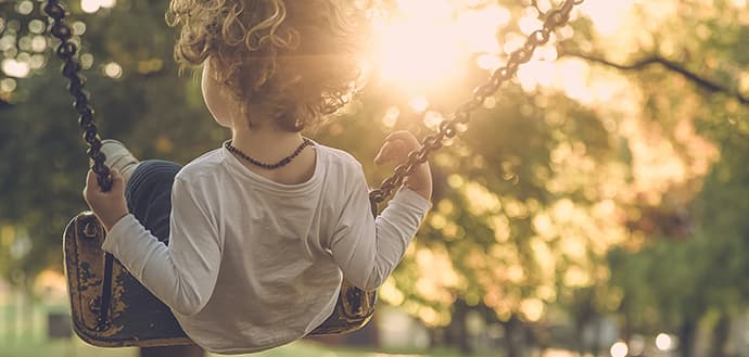 young blonde child sits on a swing with back to camera, sunlight shines through trees