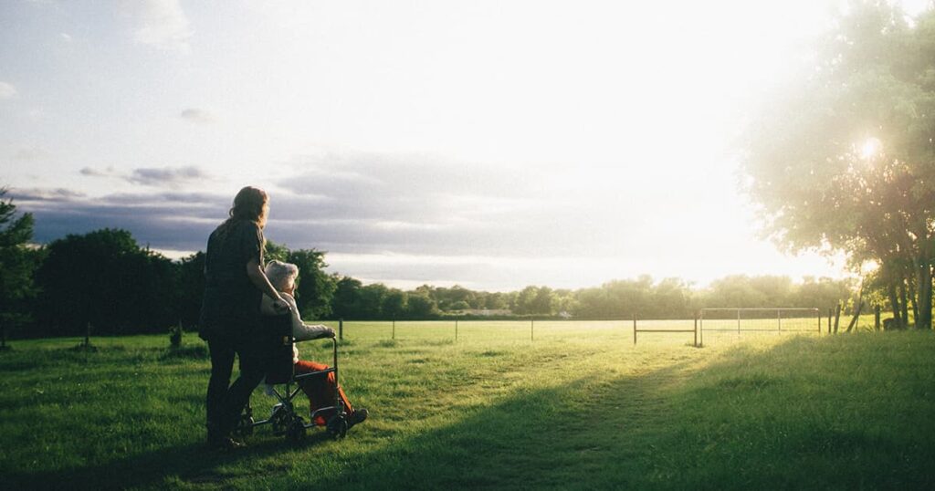 Woman pushes grandma who is in wheelchair out in a field at sunset