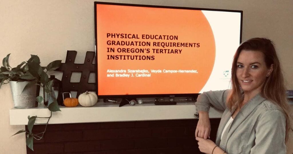 Alex Szarabajko stands in front of TV screen with the title of the team's presentation at the Western Society of Kinesiology and Wellness conference.