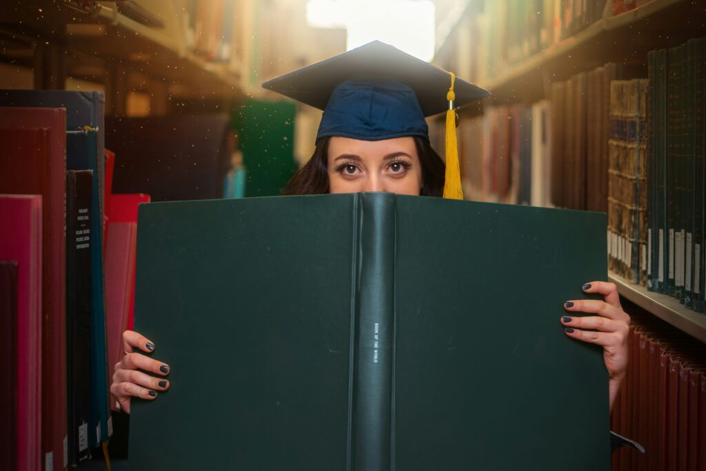 woman in a graduation cap and gown holding a massive book in front of her face at the library