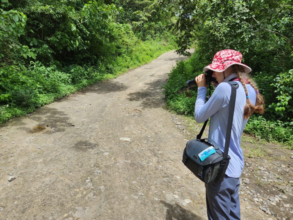 a woman in a long sleeve shirt, pants, and hate takes a photo of a butterfly sitting on a path.
