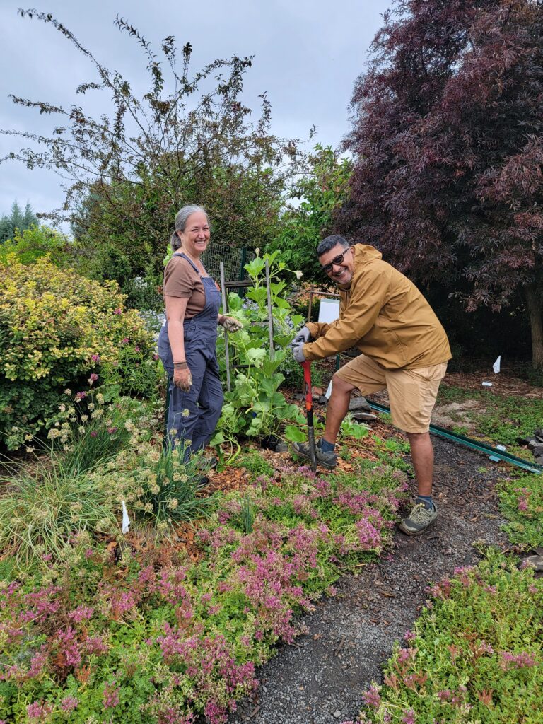 wo people working in a garden, one holding a shovel and the other standing nearby smiling, surrounded by flowering plants, shrubs, and trees on a cloudy day.