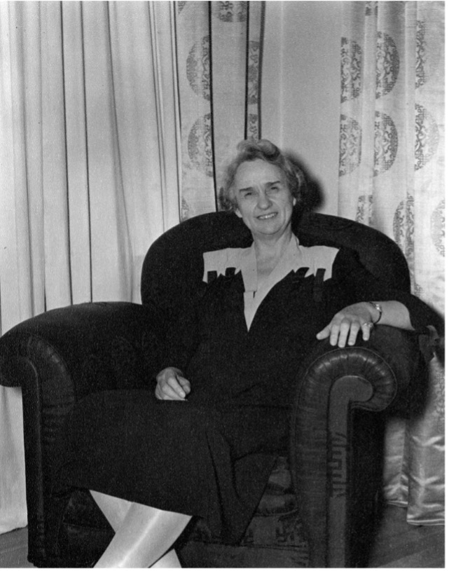 A white woman sits in a large armchair, smiling, with patterned curtains in the background.