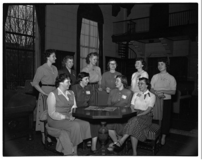 Group of nine white women around a table in a room with a large window and spiral staircase.