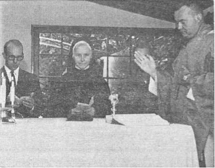 Four white individuals at a table covered with a white tablecloth in a formal setting, with windows and foliage in the background.