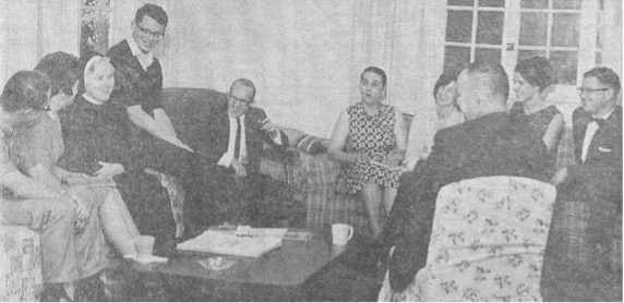 A group of white people gathered in a living room, seated around a coffee table with a newspaper and cup, in a black and white photo.