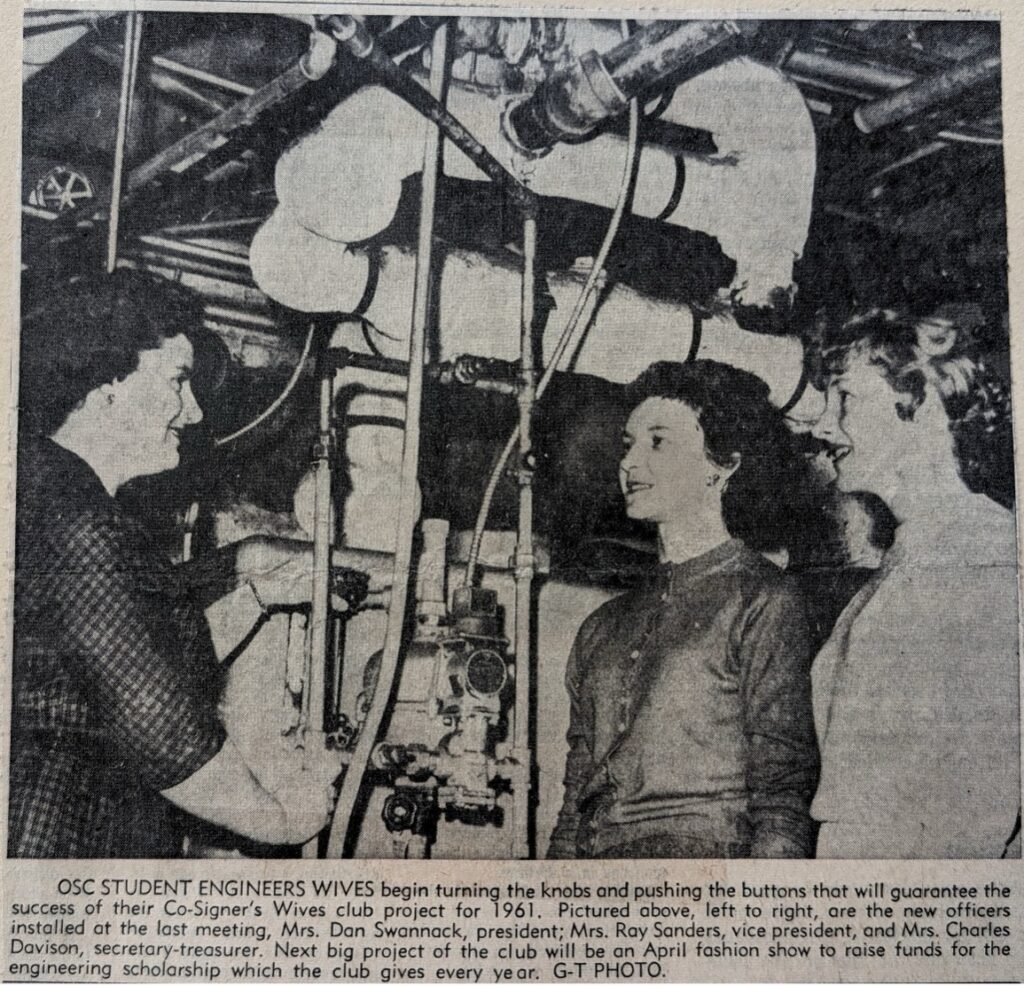 Three white women stand in front of a complex array of industrial pipes and machinery.