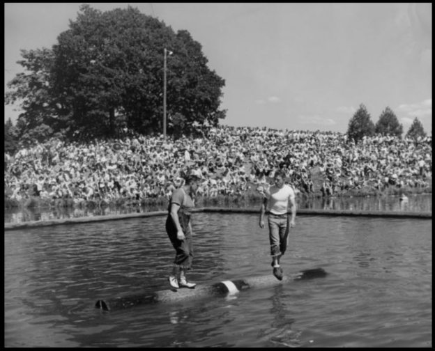 Two people standing on a log on a lake with a crowd watching.