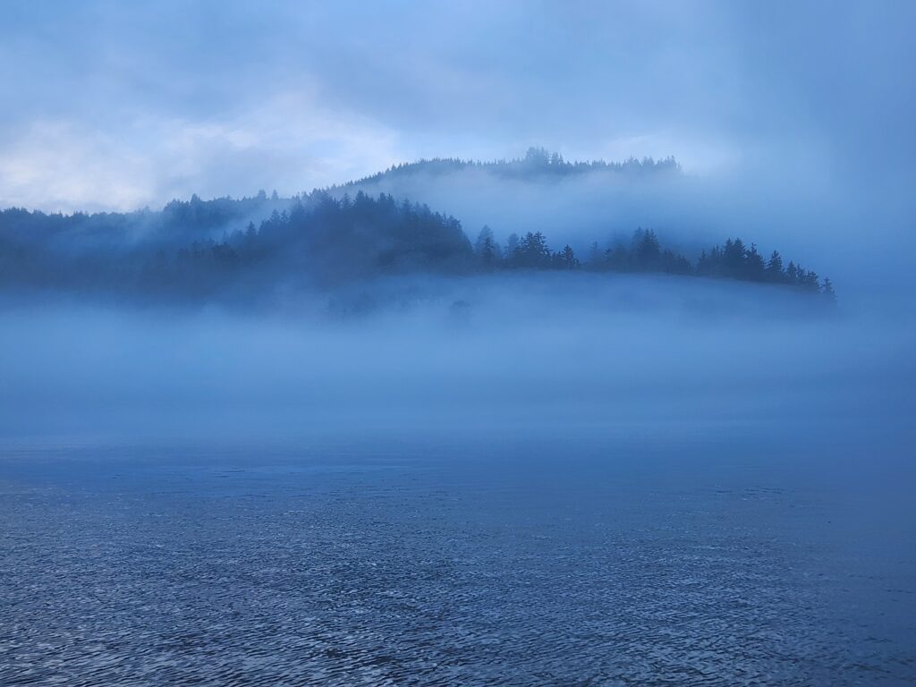 A view from on the ocean looking back towards fog rolling over the hills. Everything is blue. 