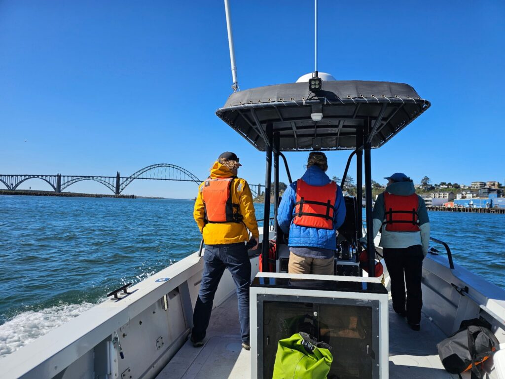 Three people in a row wearing life jackets on a boat with a center console heading towards a large arch bridge. 
