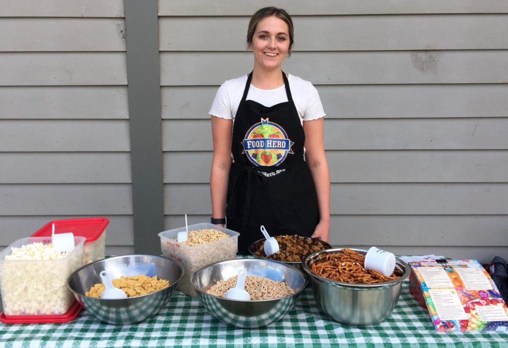 A woman in a black Food Hero apron stands at a table filled with trail mix ingredients.