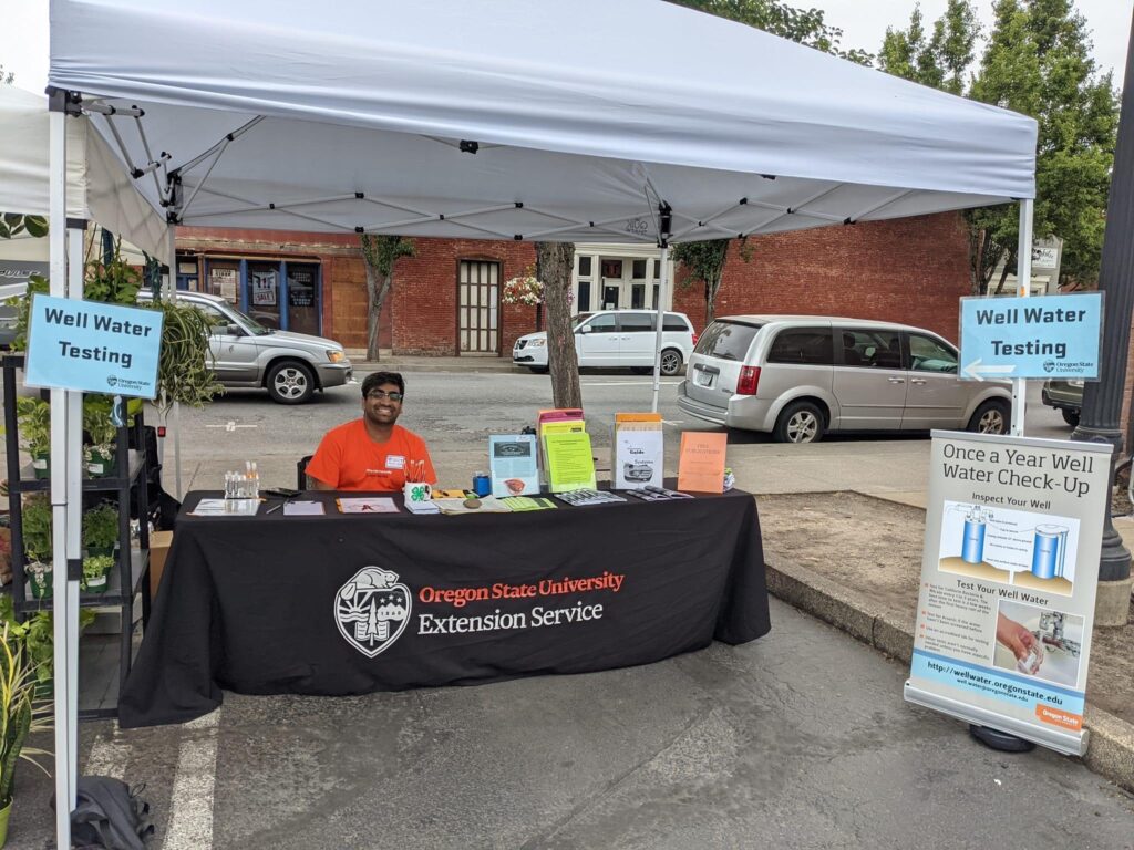 A man in an orange shirt sits under a canopy at an outdoor table, surrounded by signs and papers about well water testing.