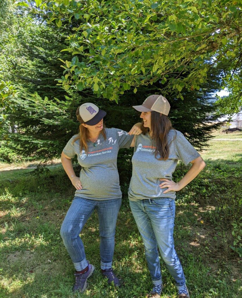 Two women in gray t-shirts and jeans stand next to each other, arm in arm, under a tree.