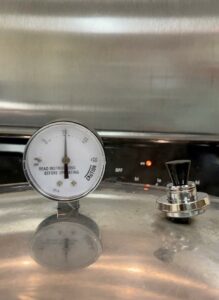 Dial gauge pressure canner at 10 pounds of pressure and a ten pound weight in a tilted position on top of the canner's vent port. This is an example of a weighted gauge being used on a dial gauge canner.