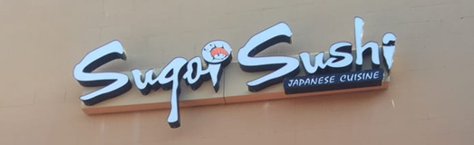 Beige building wall with a white logo that says "Sugoi Sushi" on it in bold, with "Japanese cuisine" written below in black. There is a cartoon drawing of a sushi roll above the "o" on "Sugoi".