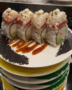 Four sushi rolls are on a stack of plates, stuffed with imitation crab and avocado, wrapped in seaweed and rice, with imitation crab, raw tuna, and a drop of kewpi mayonaise on top. The top plate is black, and the ones bellow are yellow and green.