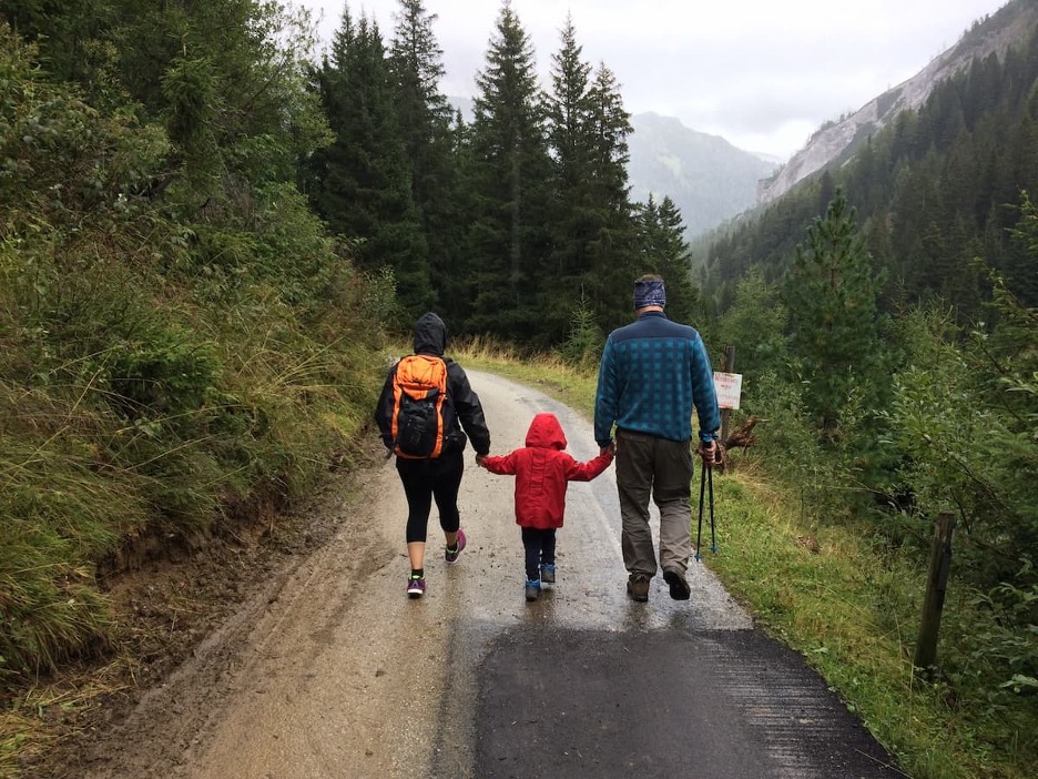 Two adults holding hands with a child walking along a path in the mountains / Dos adultos y un niño caminan de la mano por un sendero en las montañas.