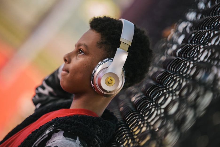 A child wearing large headphones leading against a fence