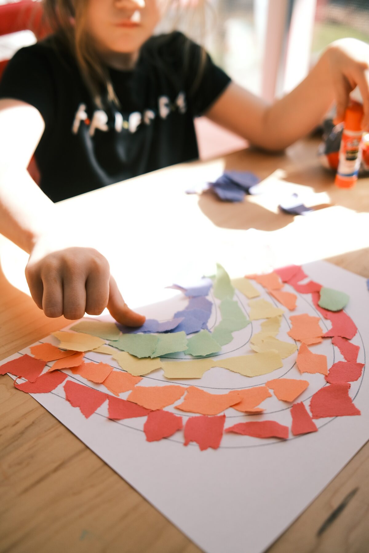 A child gluing colored scraps of paper into a rainbow shape.