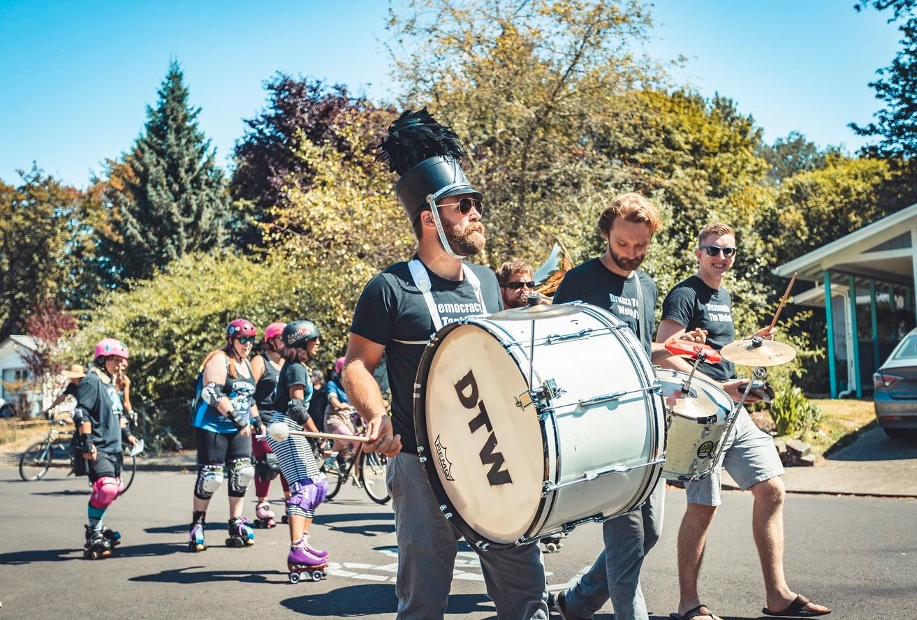 A marching band with drums and rolly derby members parade down a tree-lined Corvallis street.