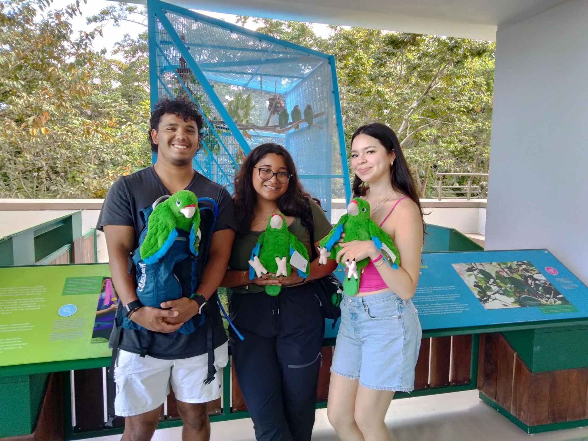 Three smiling students, holding stuffed green parrots.