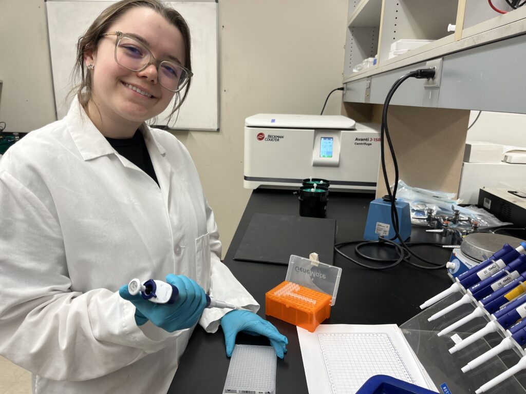 Student holds pipette in gloved hands, as they prepare to load a qPCR plate. They are looking up and smiling at the camera.