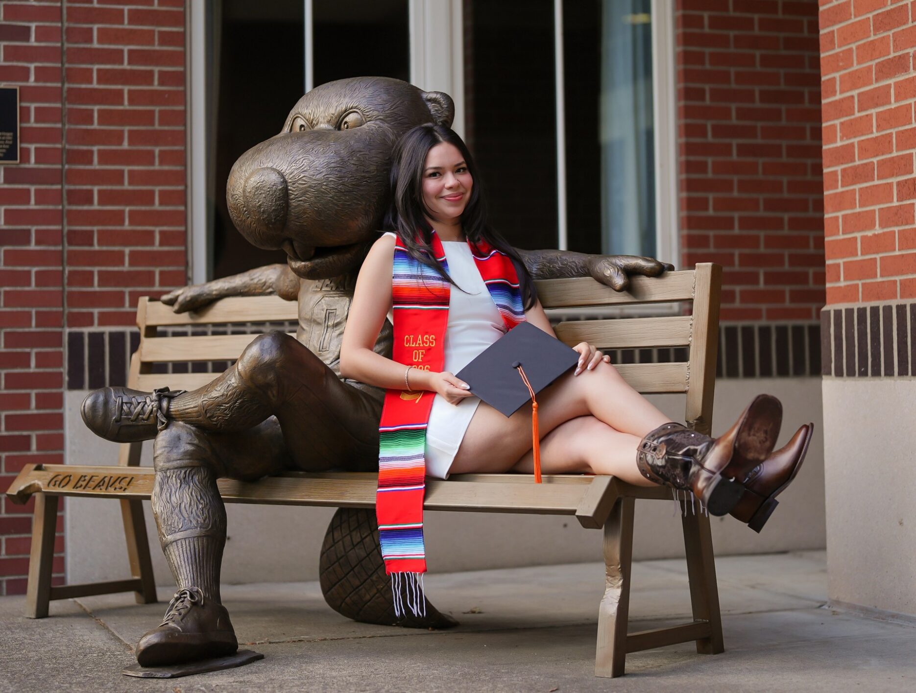 A BRR student sits on a bench next to a statue of Benny the Beaver. She is holding her graduation cap, and wearing a colorful graduation stole.