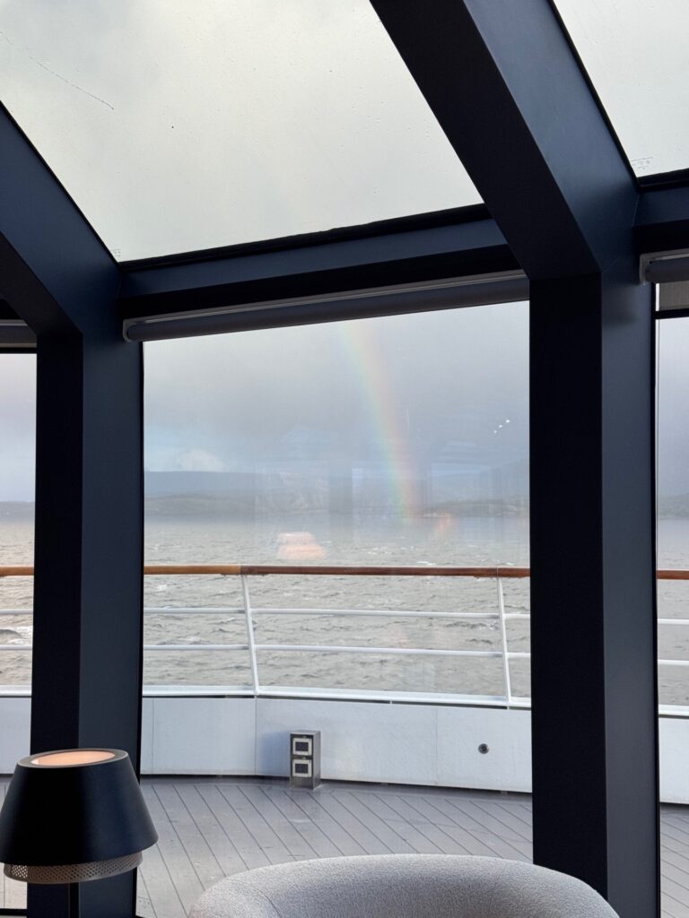 A rainbow over the water viewed through a large window from on the ship.
