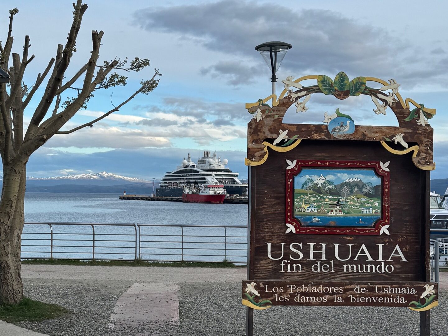 The Ushuaia sign that reads "Fin del Mundo. Los Pobladores de Ushuaia les damos la bienvenida" with the cruise vessel behind it at port in the distance and a bare tree on the left.