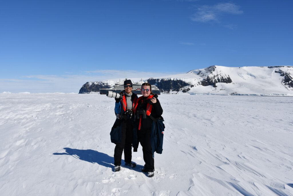 Two women smileing and holding cameras up while standing on ice with a cruise ship and large snow covered mountain in the distant background.